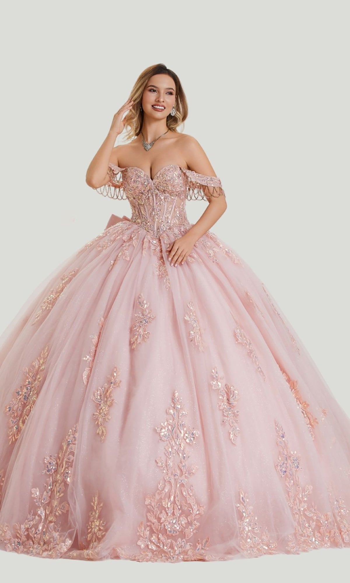 A woman wearing the Quinceanera Dress 26409 by Stella Couture in blush, featuring floral embroidery and a full tulle skirt, stands against a plain background.